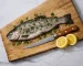 Whole seasoned fish with herbs and lemon halves on a handcrafted beech chopping board beside a wooden‑handled knife.