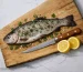 Whole seasoned fish with herbs and lemon halves on a handcrafted beech chopping board beside a wooden‑handled knife.